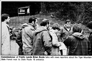 A crowd of people in rain coats stand around a man with large eyeglasses in the woods. 